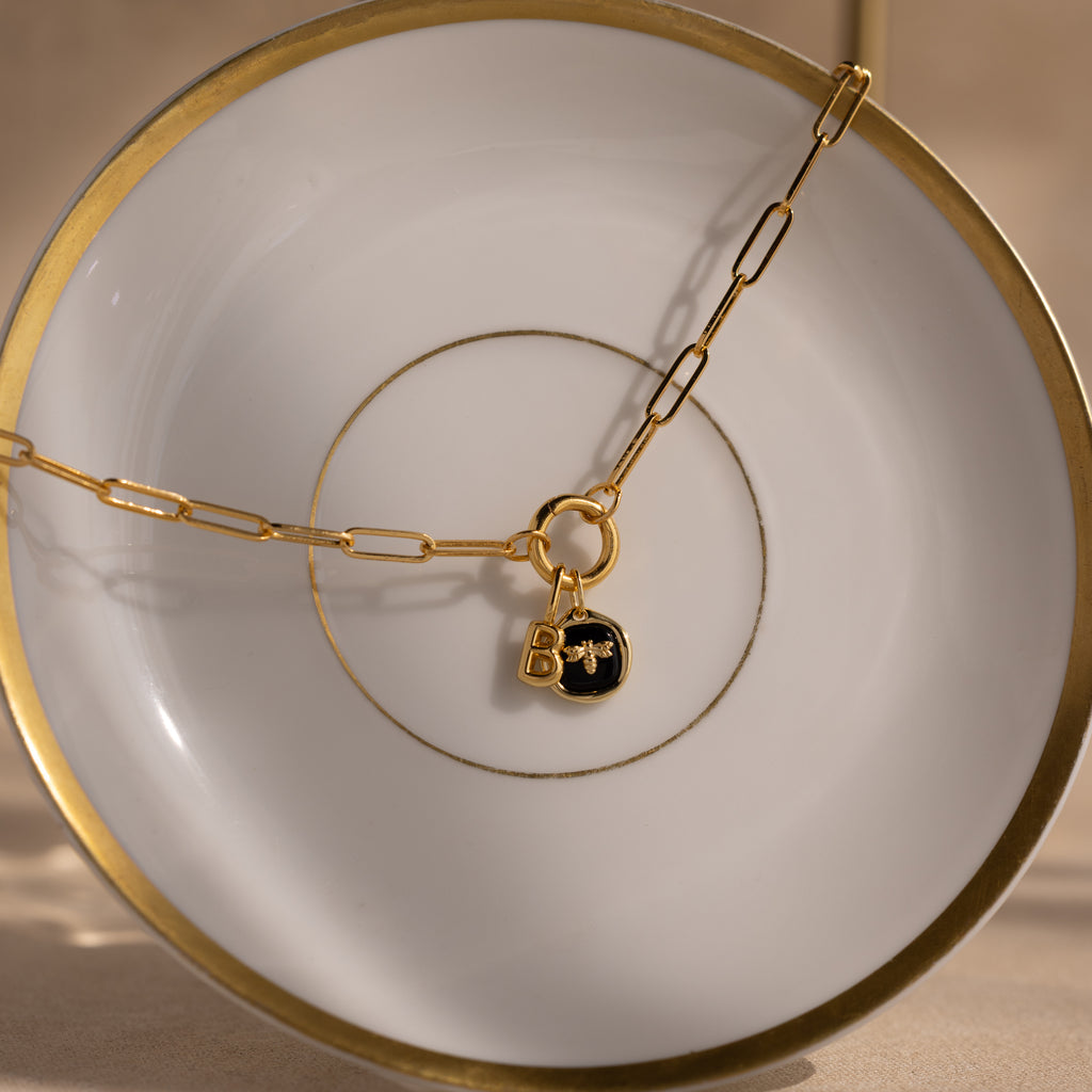 Onyx Honeybee Charm: A gold chain with a round black onyx pendant and a gleaming bee charm, elegantly draped over a white and gold-rimmed plate.