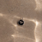 The Onyx Honeybee Charm in Silver rests on textured sand, showcased against a black background with soft sunlight.