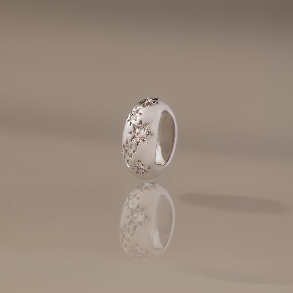 The Sprinkle Diamond Dome Charm in Silver stands upright on a reflective surface, its starburst-set stones and star-shaped engravings shining against a soft beige background.