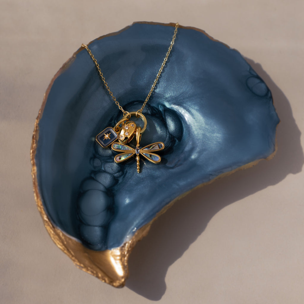 Sprinkle Diamond Dome Charm in 18K Gold featured on a gold dragonfly necklace with other charms, elegantly displayed in a blue and gold shell-shaped dish.