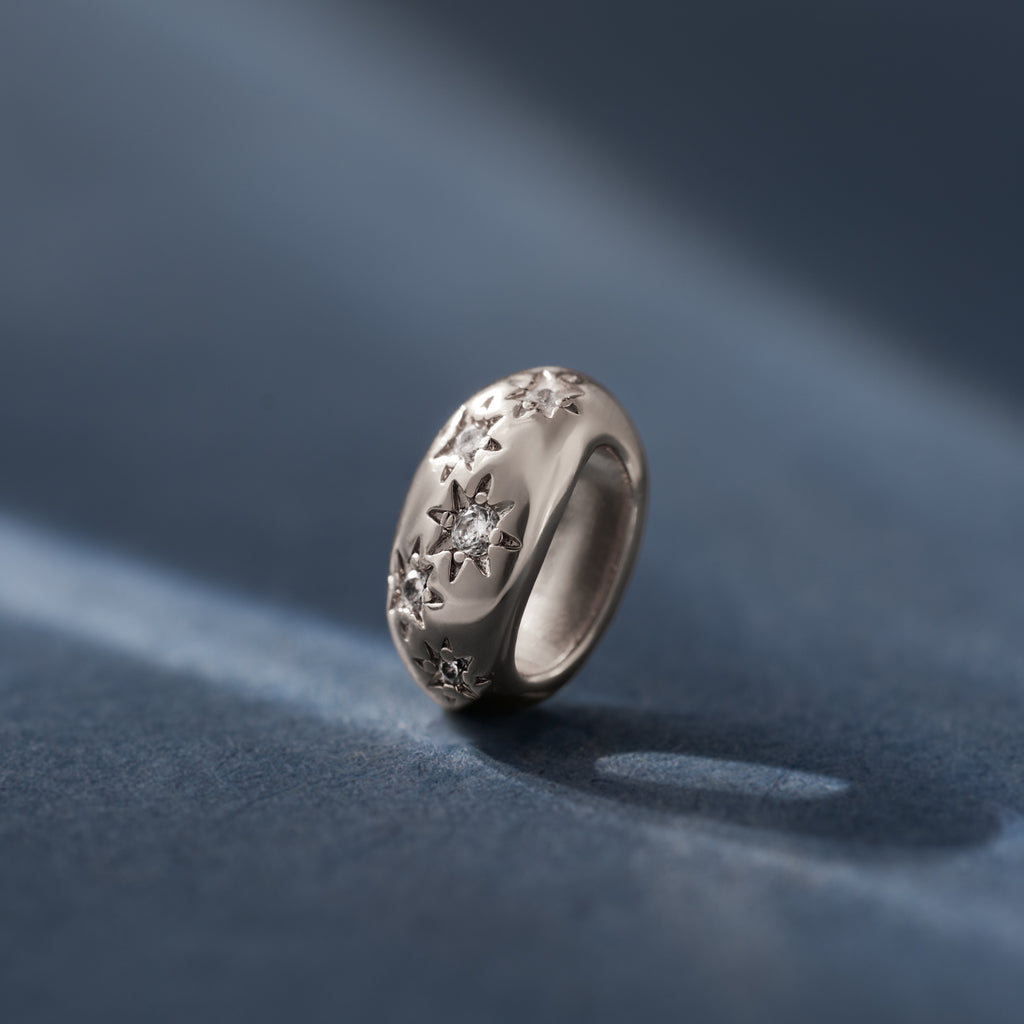 The Sprinkle Diamond Dome Charm in Silver features engraved star patterns and a central small diamond—ideal for layering in your charm stack or pairing with a diamond dome charm, shown here on a blue surface.