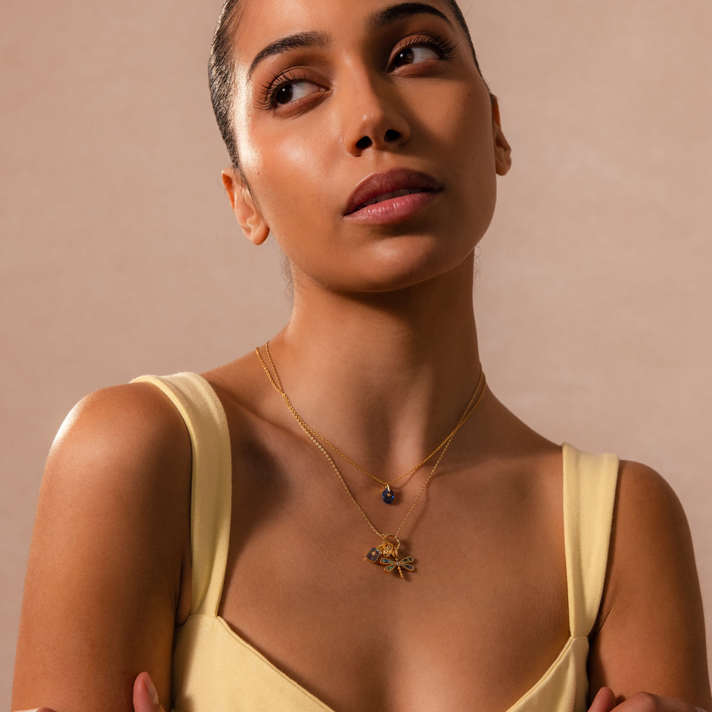 A woman in a yellow dress wears layered gold necklaces, featuring the Sprinkle Diamond Dome Charm in 18K Gold from her charm stack, as she gazes upward against a soft beige background.