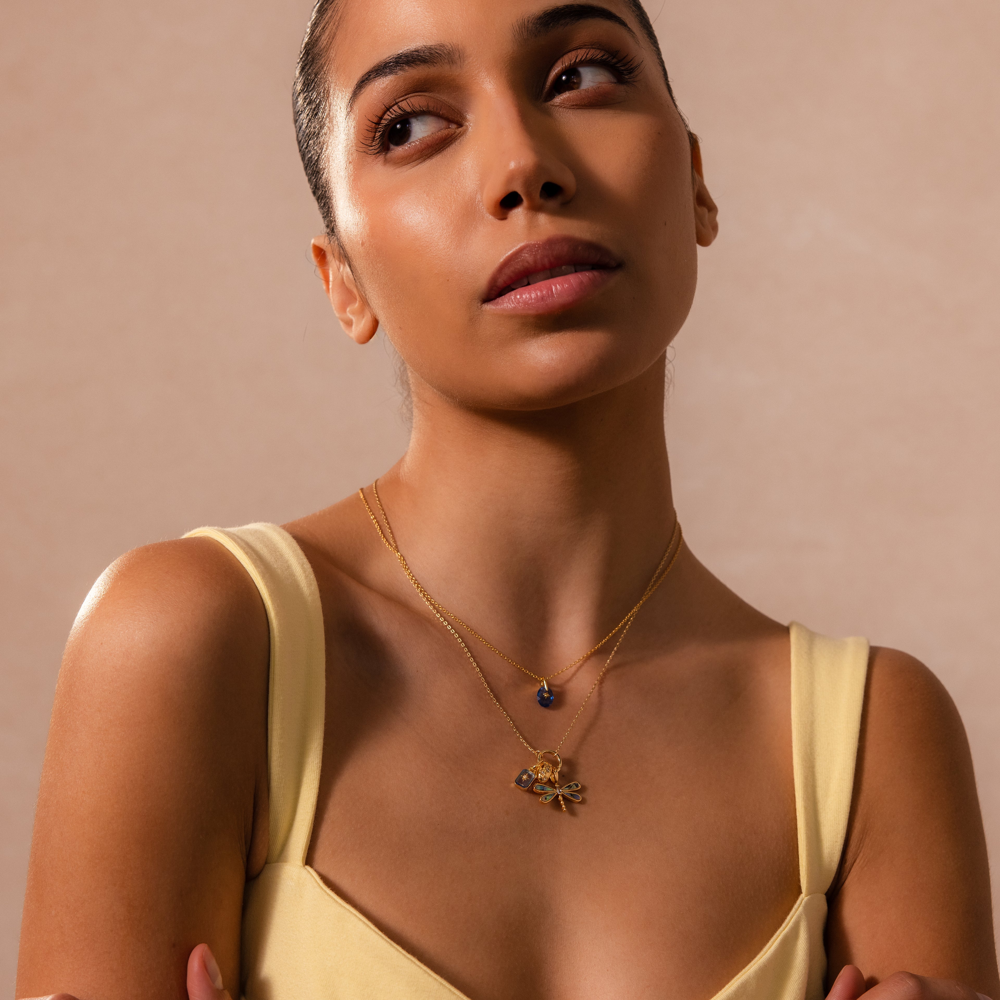 A woman in a yellow dress wears layered gold necklaces, featuring the Sprinkle Diamond Dome Charm in 18K Gold from her charm stack, as she gazes upward against a soft beige background.