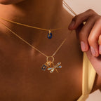 A person wears two gold necklaces: one with a blue charm and another with the Abalone Dragonfly Charm, which features iridescent wing details and nature-inspired accents.
