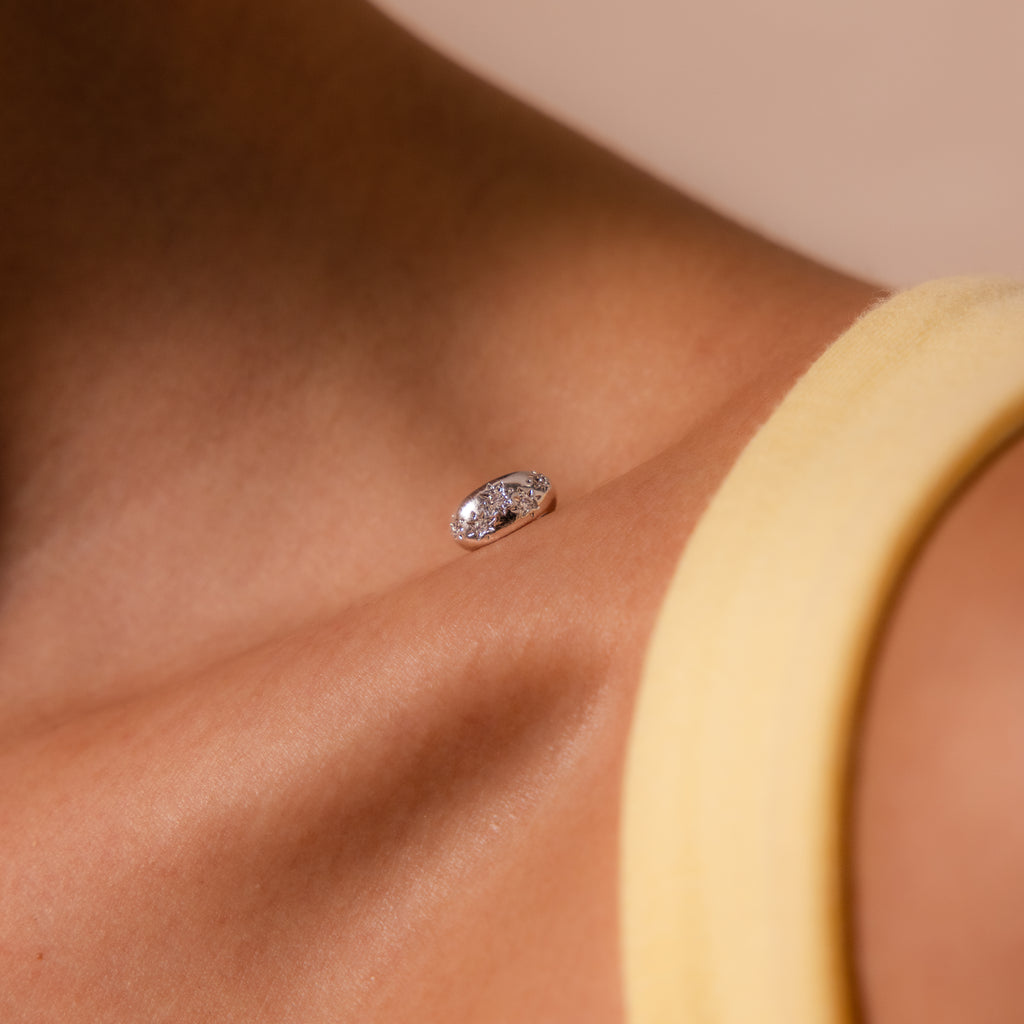 A close-up of a dermal piercing with a jeweled stud on the collarbone, beside a yellow strap and complemented by a delicate stack featuring the Sprinkle Diamond Dome Charm in Silver.