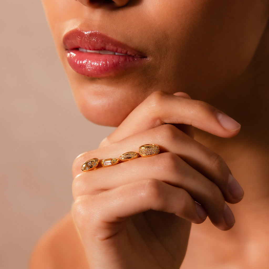 Close-up of a woman’s lips and hand, wearing a gold ring with starburst-set stones and the Sprinkle Diamond Dome Charm in 18K Gold on her charm stack.