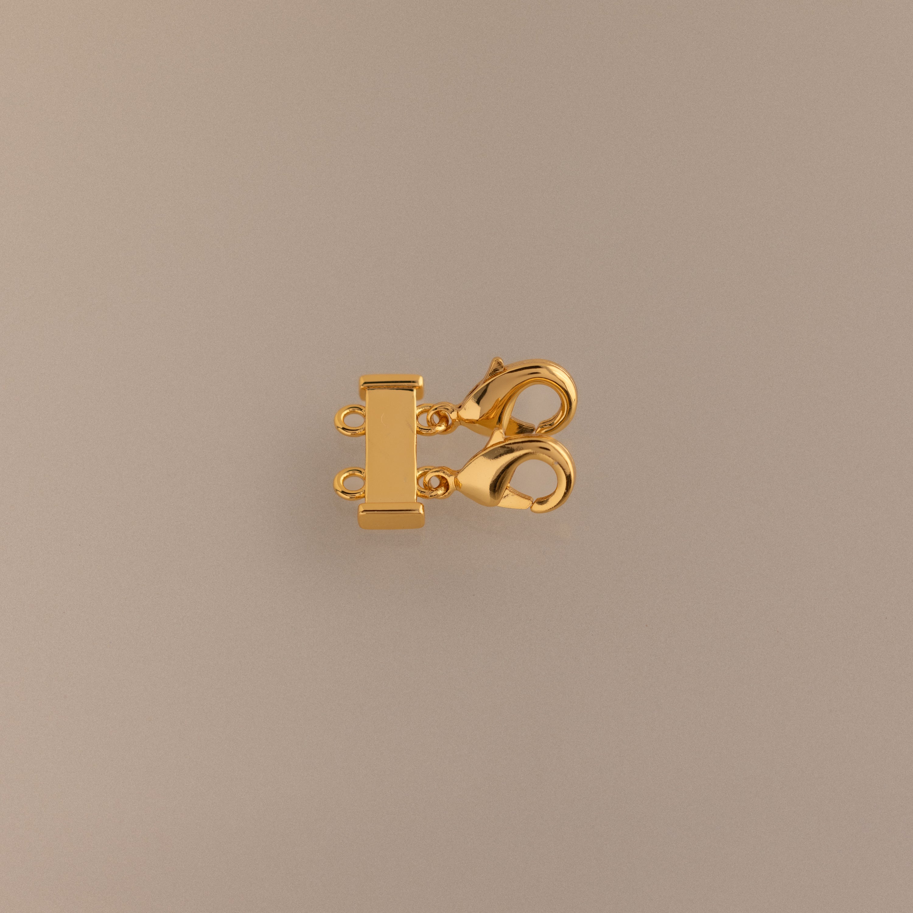 The Double Layering Clasp in 18K Gold features a triple-strand design with two lobster claw closures, ideal for layering necklaces. Displayed on a neutral beige background.