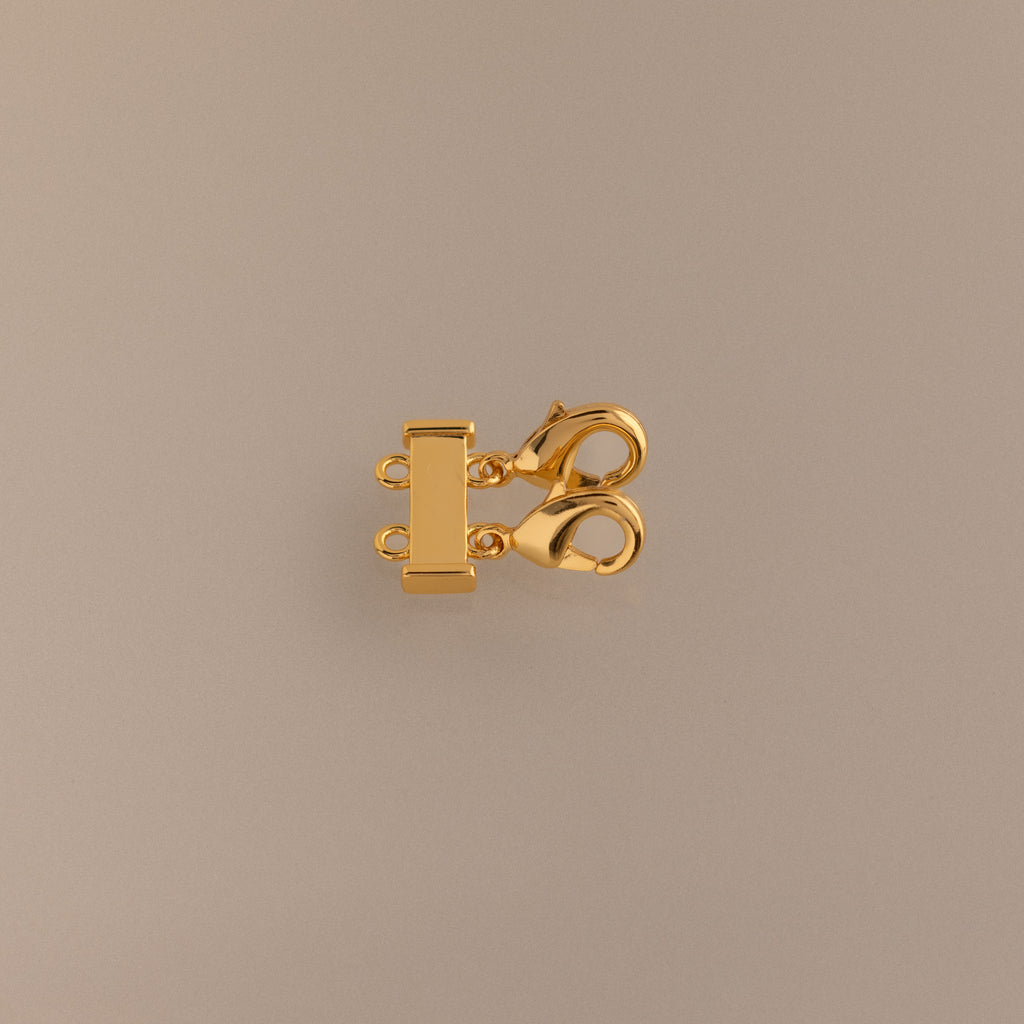 The Double Layering Clasp is a gold multi-strand jewelry clasp with two lobster clasps and three connector rings, perfect for necklace layering and preventing tangling—shown on a beige background.