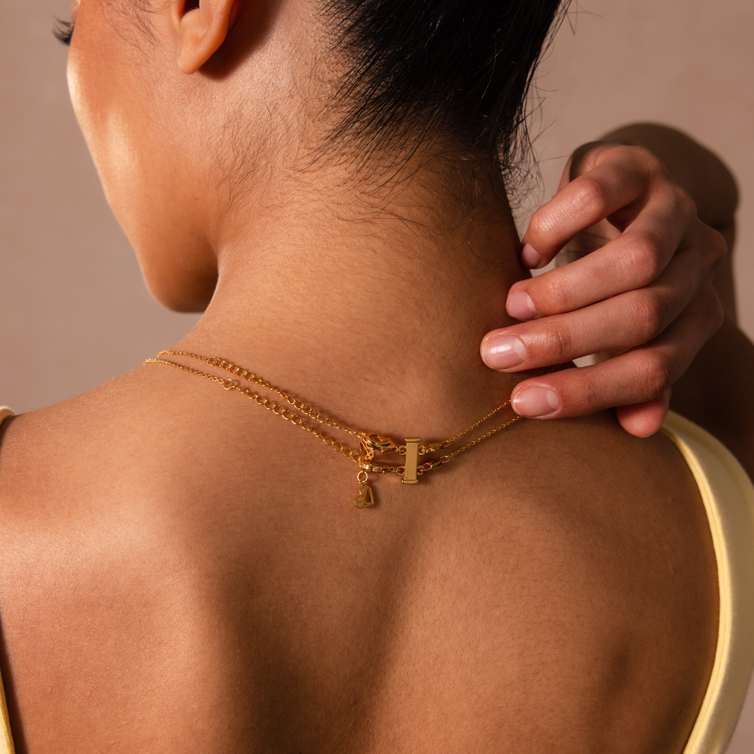 Woman touching her neck, elegantly layering gold necklaces with the Double Layering Clasp in 18K Gold visible at the back—a chic accessory for any look.