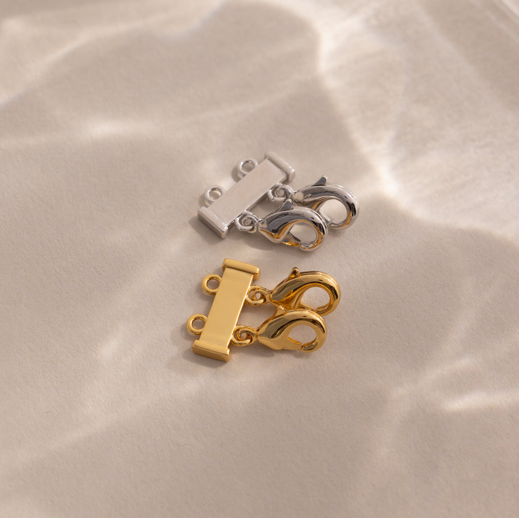 The Double Layering Clasp in 18K Gold, shown here in both silver and gold on a beige surface with soft light reflections, is perfect for effortless necklace layering.