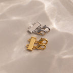 The Double Layering Clasp in 18K Gold, shown here in both silver and gold on a beige surface with soft light reflections, is perfect for effortless necklace layering.