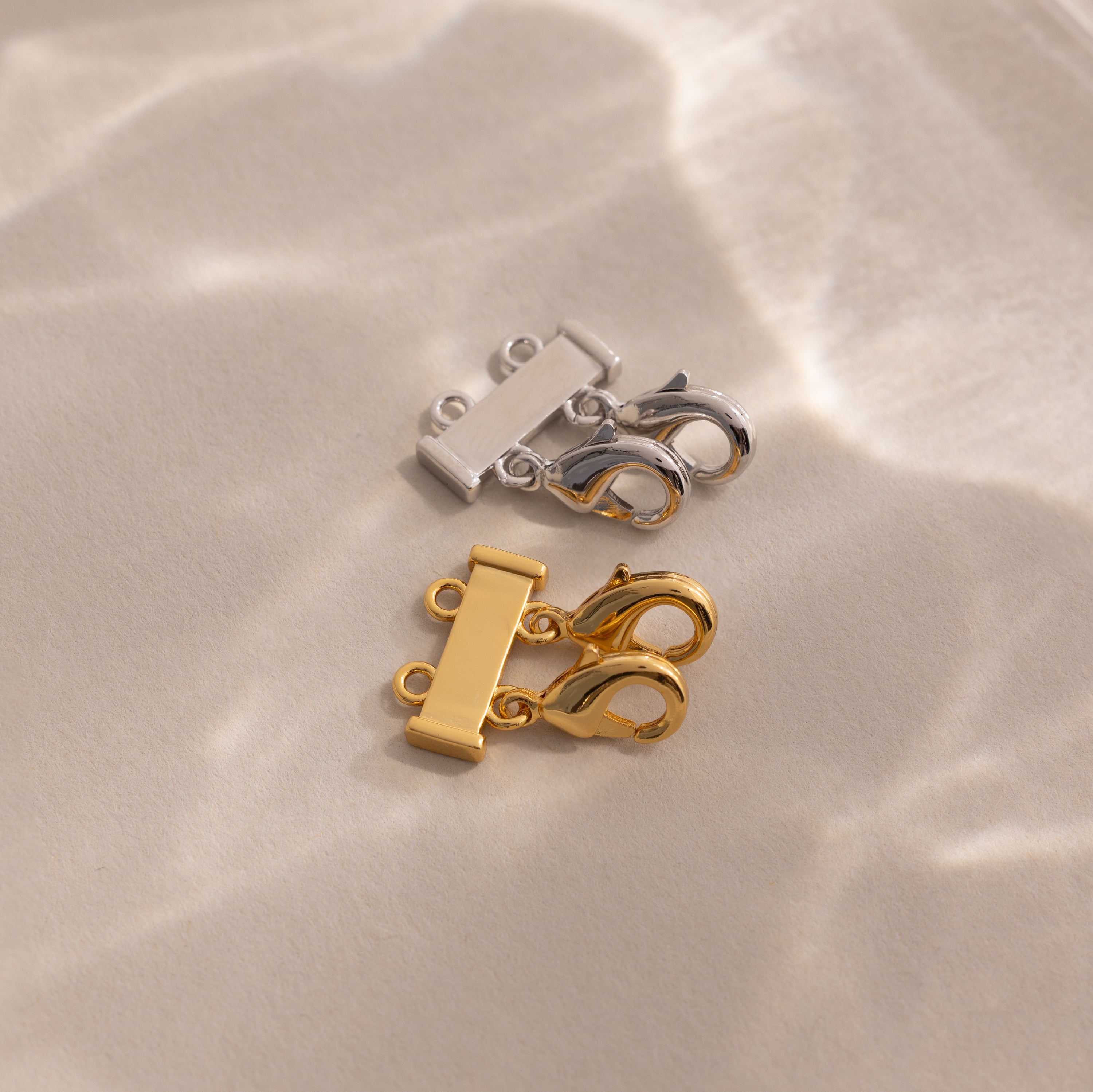 The Double Layering Clasp in 18K Gold, shown here in both silver and gold on a beige surface with soft light reflections, is perfect for effortless necklace layering.