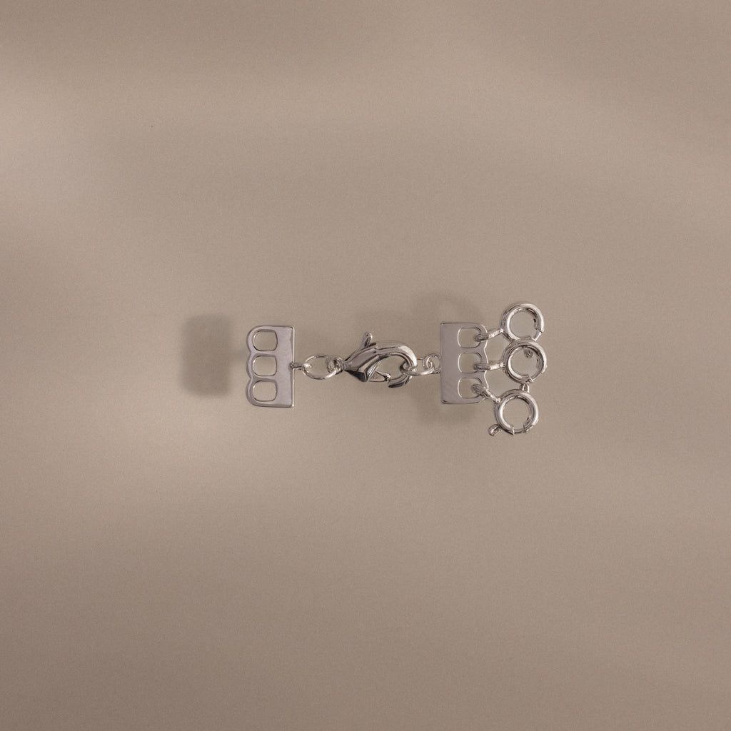 The Triple Layering Clasp in Silver features three loops and a clasp for tangle-free necklace layering, shown on a beige background.