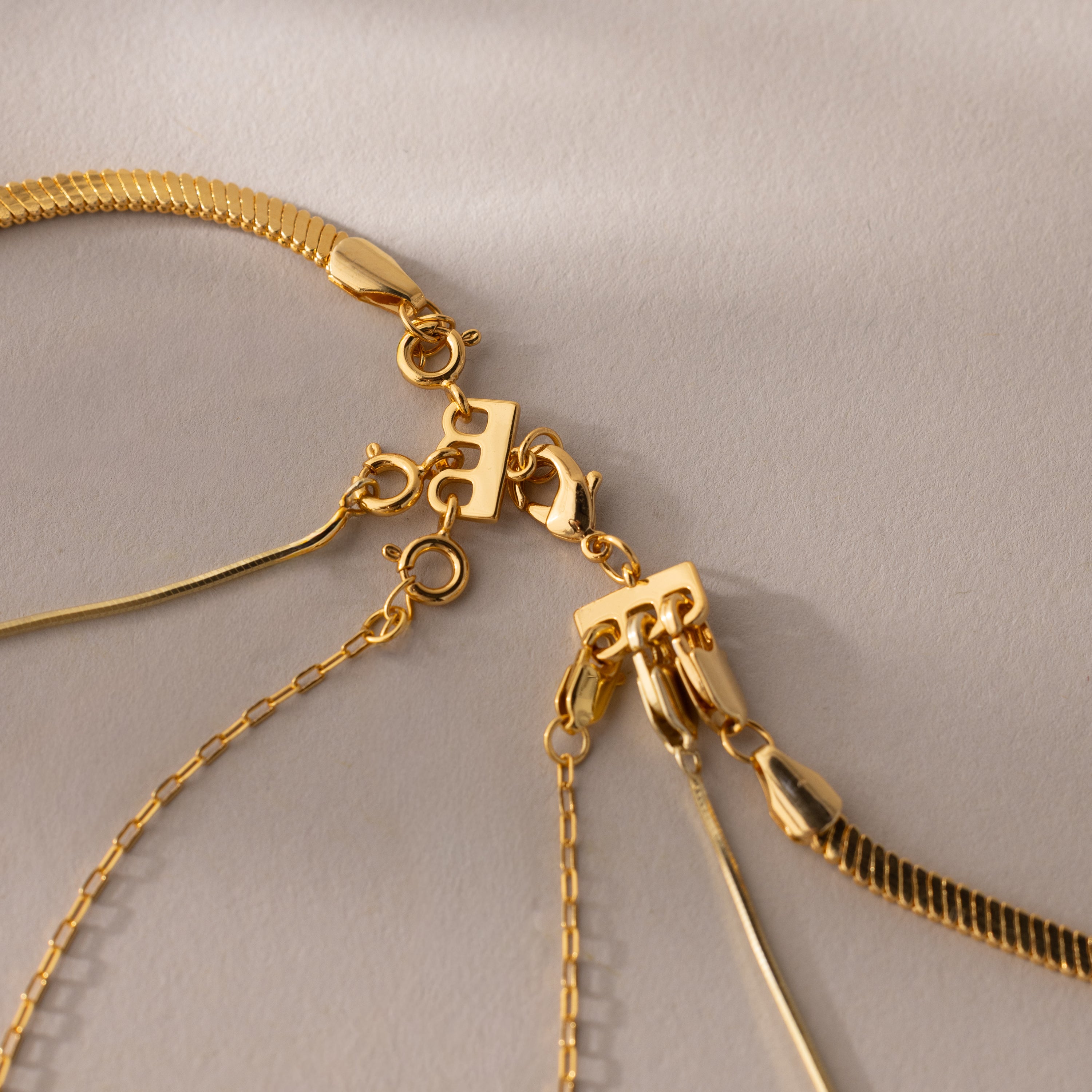 Three gold chains with unique patterns rest on a beige surface, ideal for layering. The Triple Layering Clasp in 18K Gold allows you to effortlessly mix and style gold and silver necklaces together.