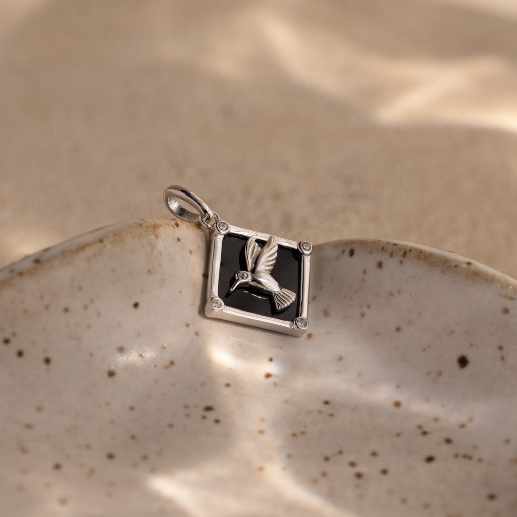 An Onyx Hummingbird Charm in Silver rests on a speckled ceramic dish with a sandy background.