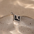 An Onyx Hummingbird Charm in Silver rests on a speckled ceramic dish with a sandy background.