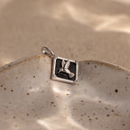 An Onyx Hummingbird Charm in Silver rests on a speckled ceramic dish with a sandy background.