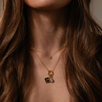 A woman with long brown hair wears the Onyx Hummingbird Charm layered with gold necklaces, creating a delicate and elegant look perfect for any occasion.
