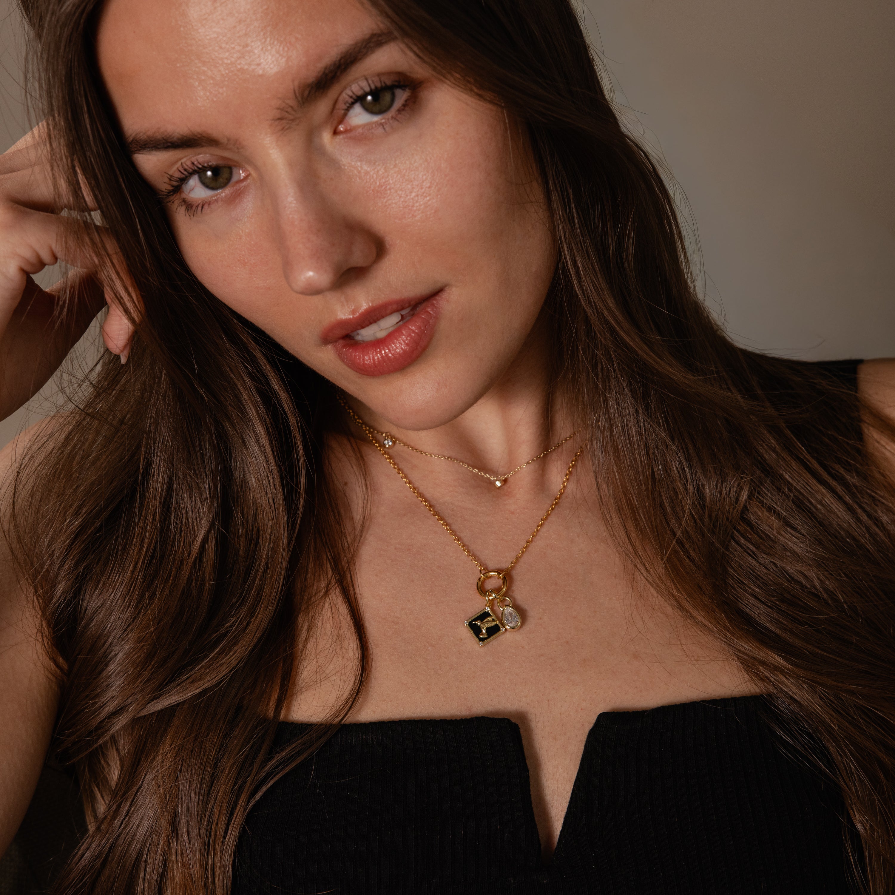 A woman with long brown hair, wearing a black top and layered gold necklaces, confidently showcases the Onyx Hummingbird Charm in 18K Gold—elegant jewelry perfect for any occasion.