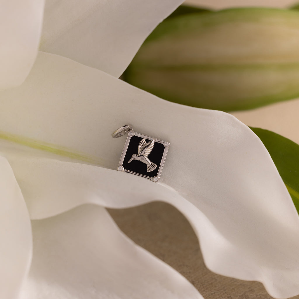 The Onyx Hummingbird Charm in Silver rests on a black square atop soft white flower petals and green leaves—a refined piece to elevate your jewelry collection.