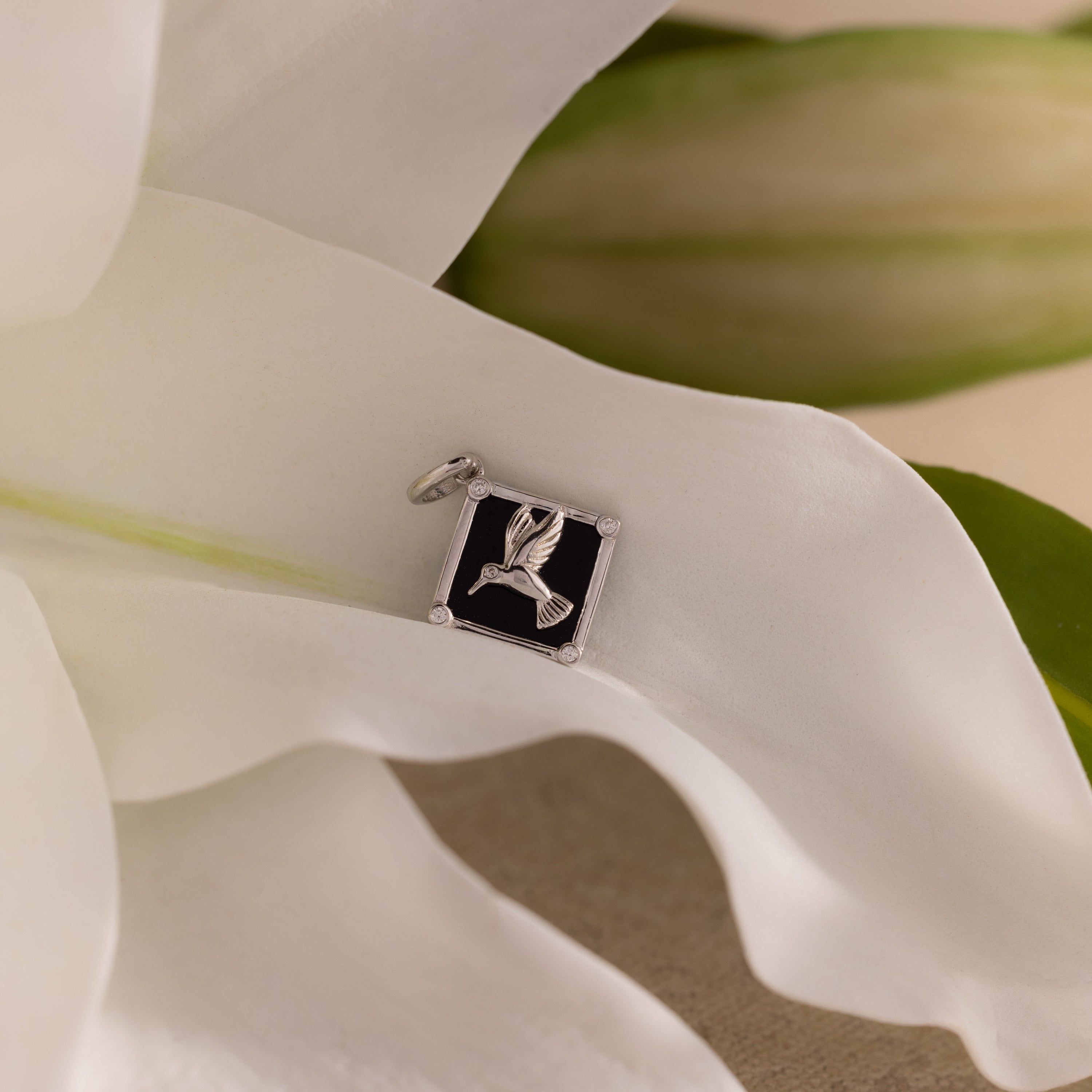 The Onyx Hummingbird Charm, a silver bird pendant, rests on a white flower with green leaves in the background.