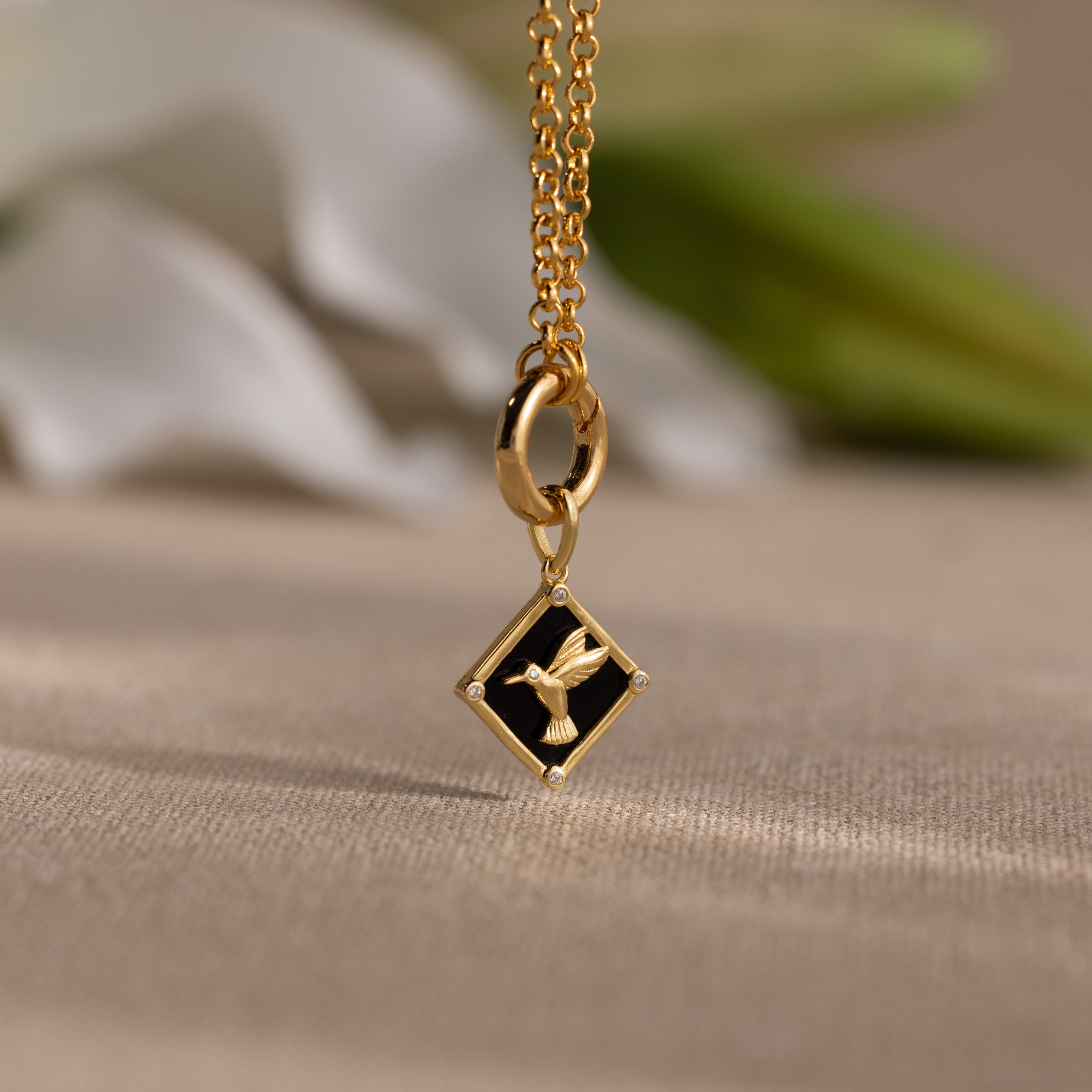 The Onyx Hummingbird Charm is a gold necklace with a square black pendant featuring a gold bird design, elegantly displayed on a beige surface.