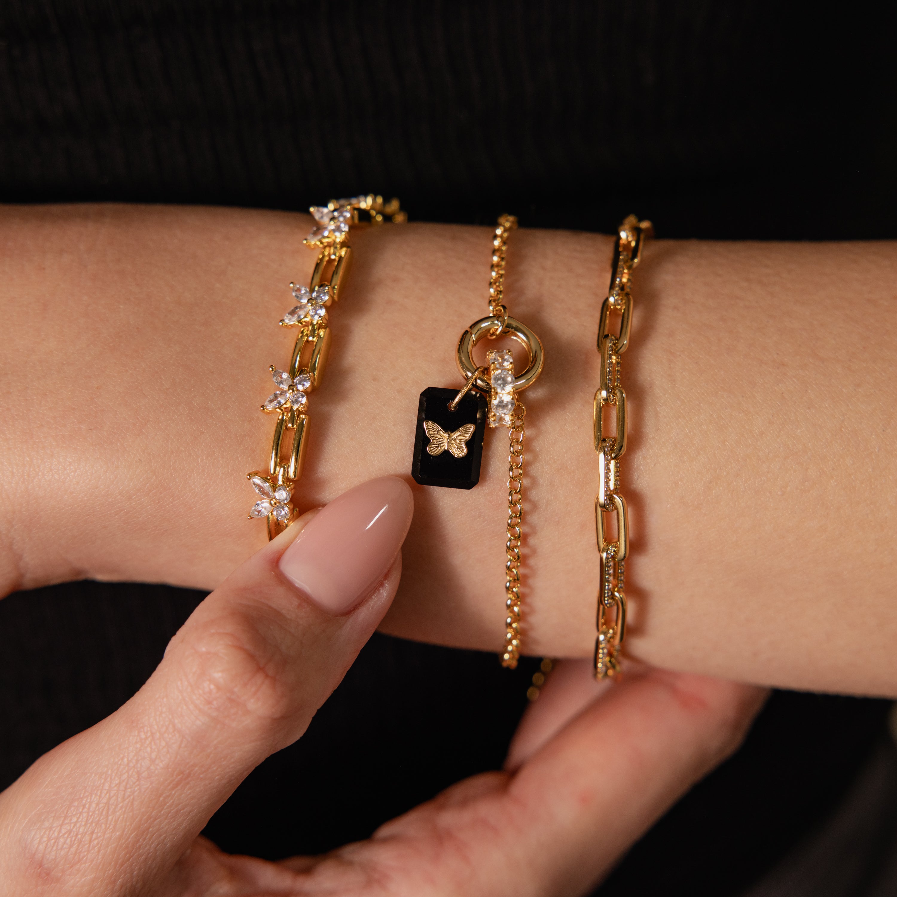 A hand adorned with three gold bracelets, including the Onyx Butterfly Charm, is elegantly displayed against a black background—ideal for those seeking unique and sophisticated jewelry.