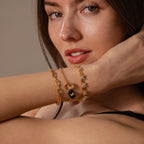 A woman wearing a black strap top raises her arm near her face, showcasing gold bracelets adorned with the Onyx Butterfly Charm.