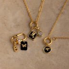 The Onyx Honeybee Charm in 18K Gold shines among three gold necklaces adorned with delicate letter, butterfly, bee, and gemstone accents, elegantly displayed on a stone surface—perfect for stylish layering or gifting.