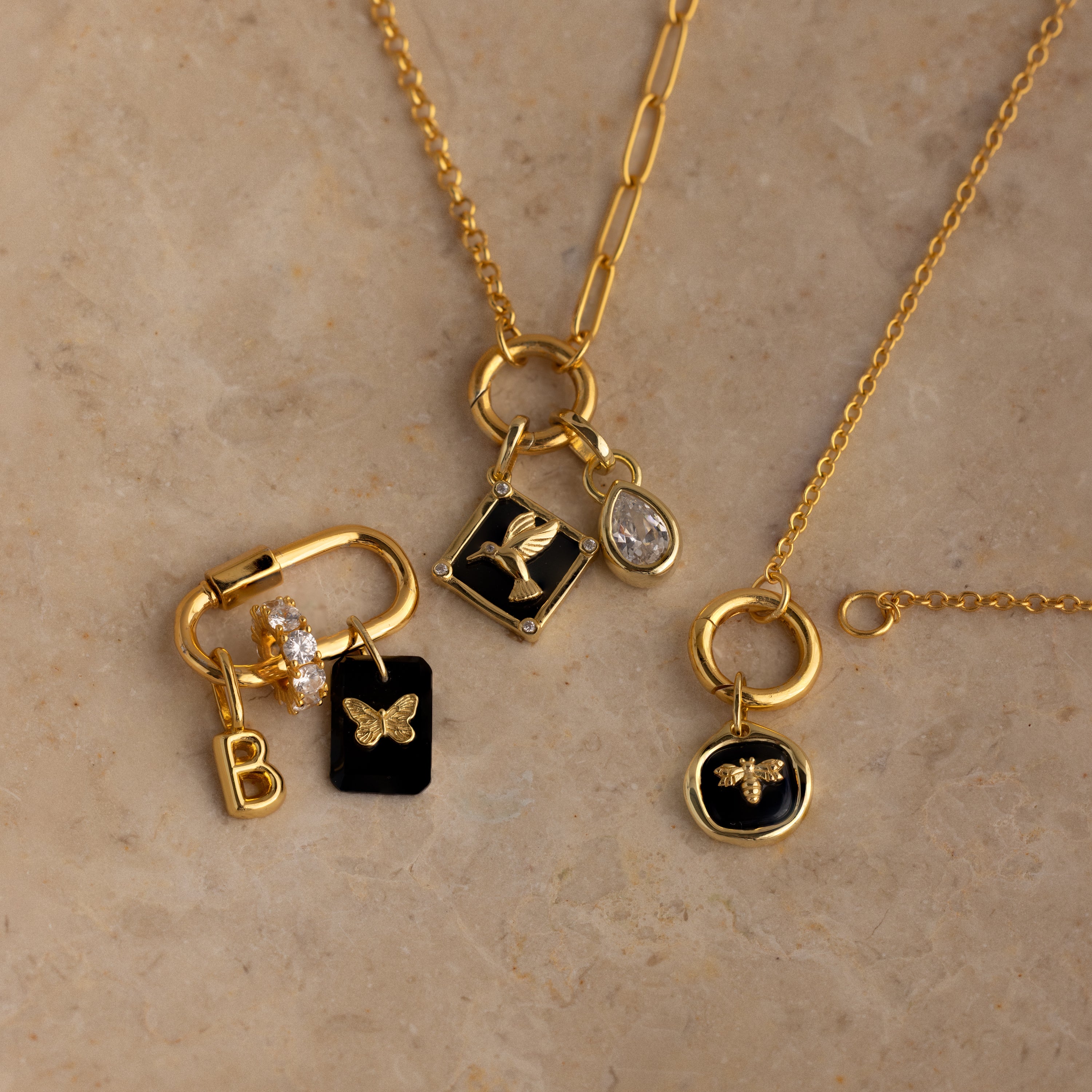 The Onyx Honeybee Charm in 18K Gold shines among three gold necklaces adorned with delicate letter, butterfly, bee, and gemstone accents, elegantly displayed on a stone surface—perfect for stylish layering or gifting.