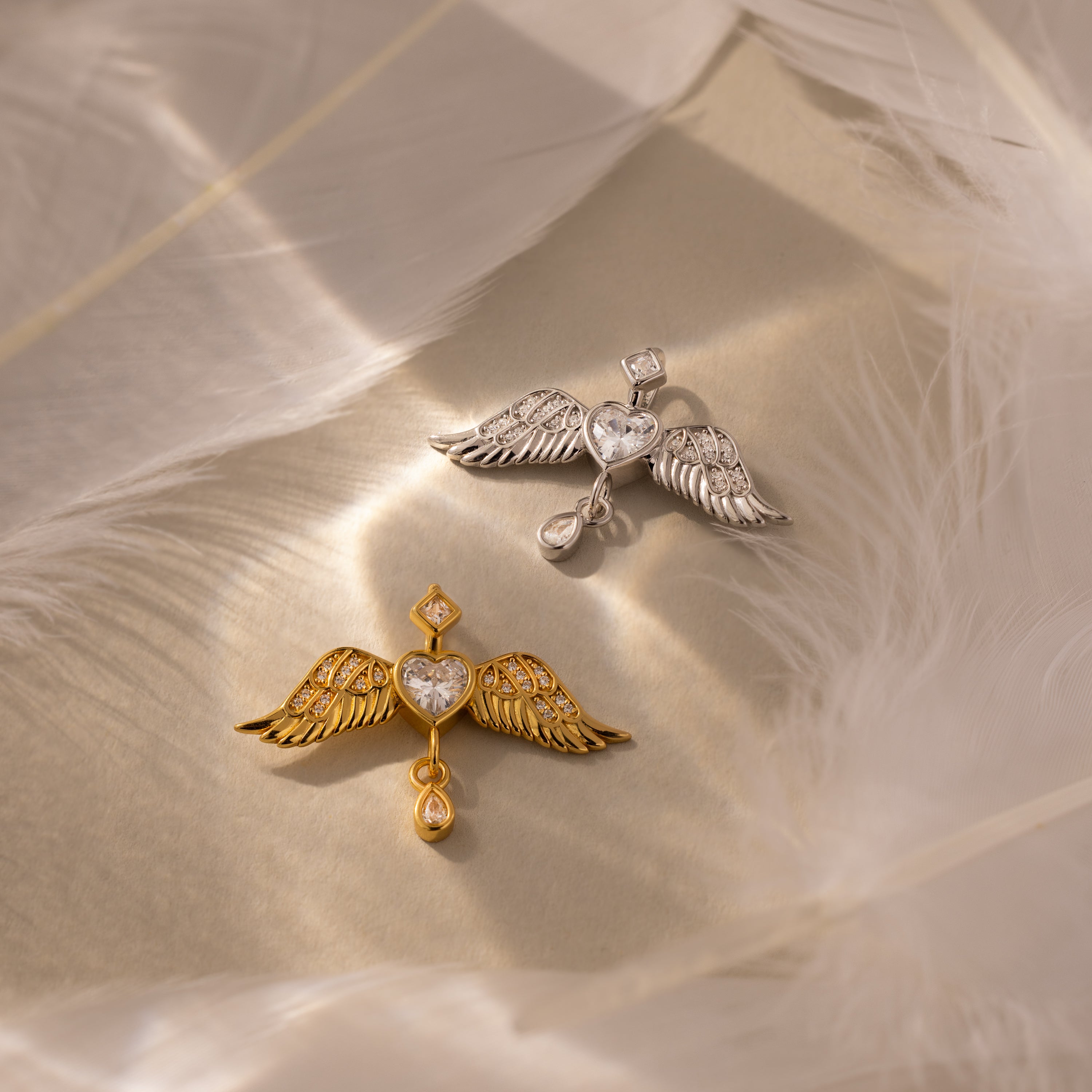 Two Angel Wing Charms—one gold, one silver—each with a heart-shaped center, rest on a feathered surface. These pendants are ideal as protection jewelry or a meaningful gift for someone special.