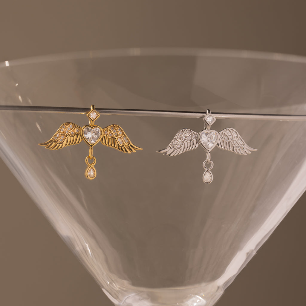 Gold and silver Angel Wing Charms with pavé diamond crystals, draped on a martini glass—ideal as protection jewelry or a sophisticated pendant.