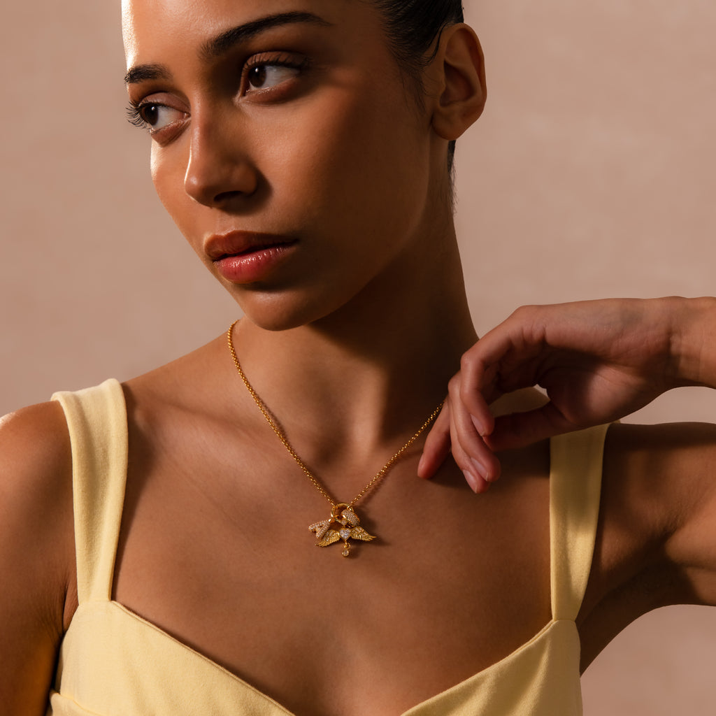A woman in a yellow top wears a gold necklace featuring the Angel Wing Charm, glancing to the side and adding a hint of protection jewelry to her elegant look.