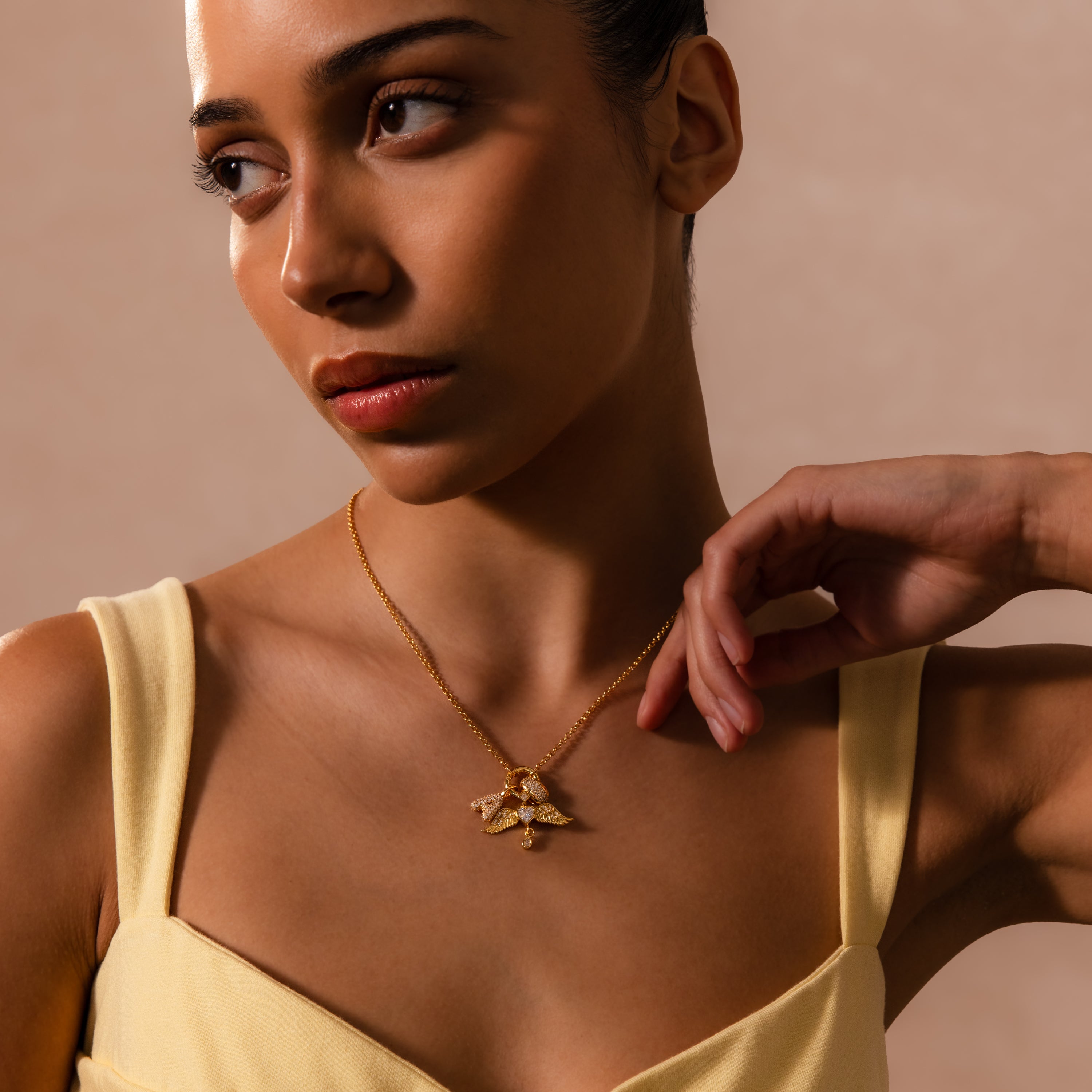 A woman in a yellow top wears a gold necklace featuring the Angel Wing Charm, glancing to the side and adding a hint of protection jewelry to her elegant look.