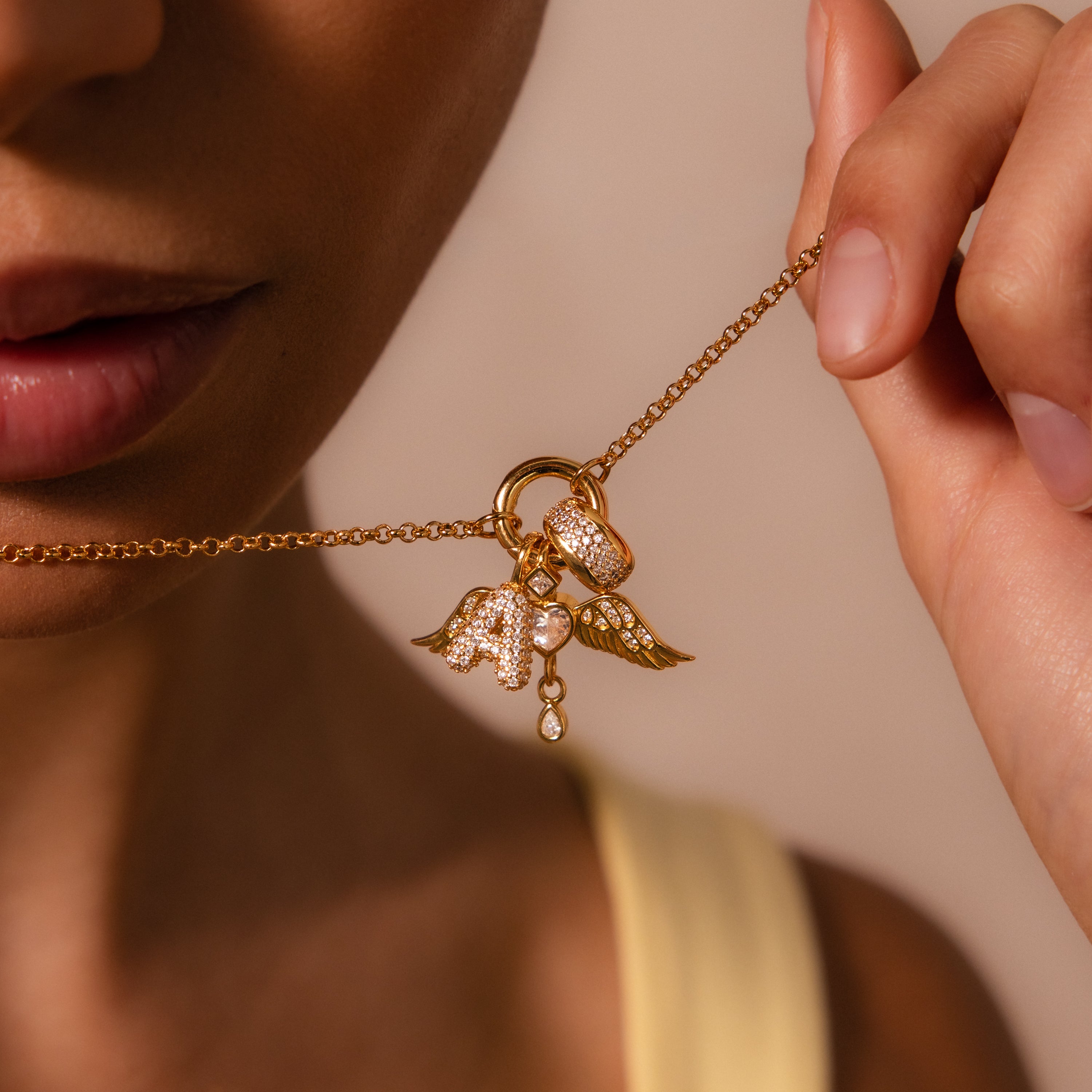 Close-up of a woman holding a Bold Pave Dome Charm gold necklace, personalized with wing and letter A charms, detailed with pearls and crystals.
