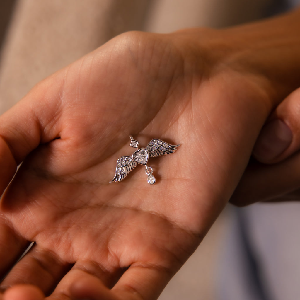 A hand holds the Angel Wing Charm in Silver, showcasing pavé-set wings shaped into a heart with two delicately dangling drop diamonds below.