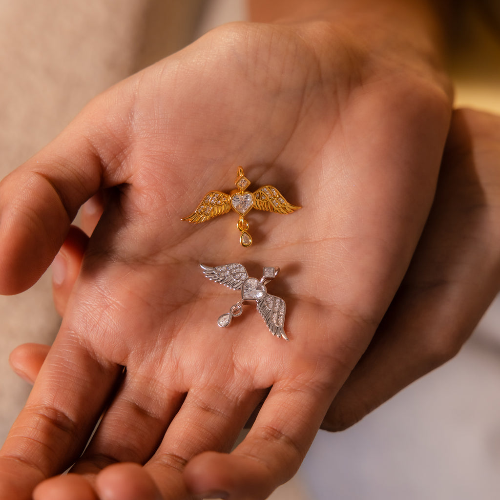 A hand holds two Angel Wing Charms—one gold, one silver—each featuring a heart-shaped center; these pendants are delicate protection jewelry.