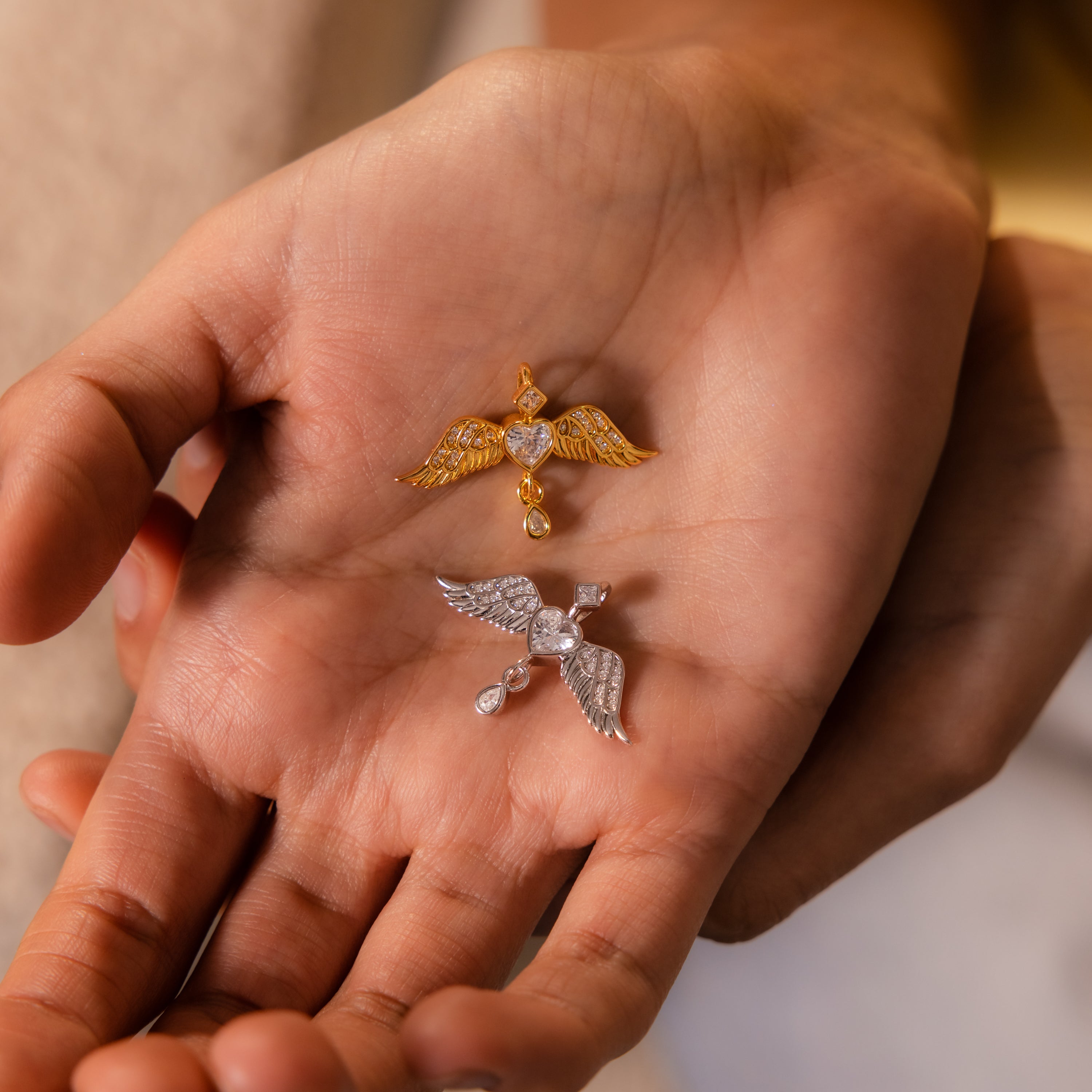 A hand holds two Angel Wing Charms—one gold, one silver—each featuring a heart-shaped center; these pendants are delicate protection jewelry.