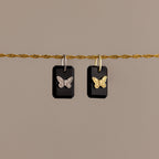Two black rectangular Onyx Butterfly Charms with butterfly designs hang from a twisted gold rope against a beige background.
