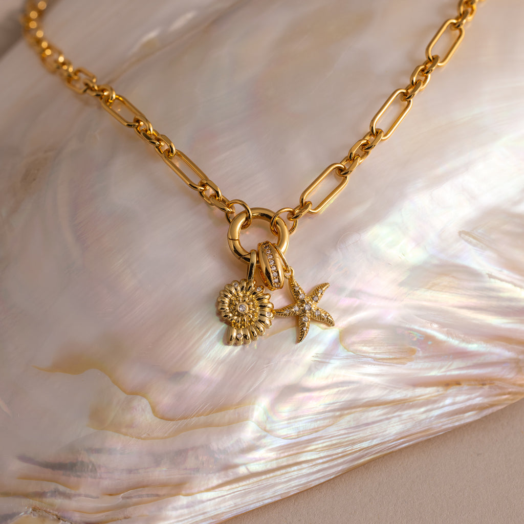 The Rolo Circle Figaro Necklace in 18K Gold features daisy and starfish charms, a statement front clasp, and is displayed on an iridescent shell for a modern update to the classic link style.