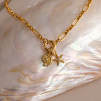 The Rolo Circle Figaro Necklace in 18K Gold features daisy and starfish charms, a statement front clasp, and is displayed on an iridescent shell for a modern update to the classic link style.