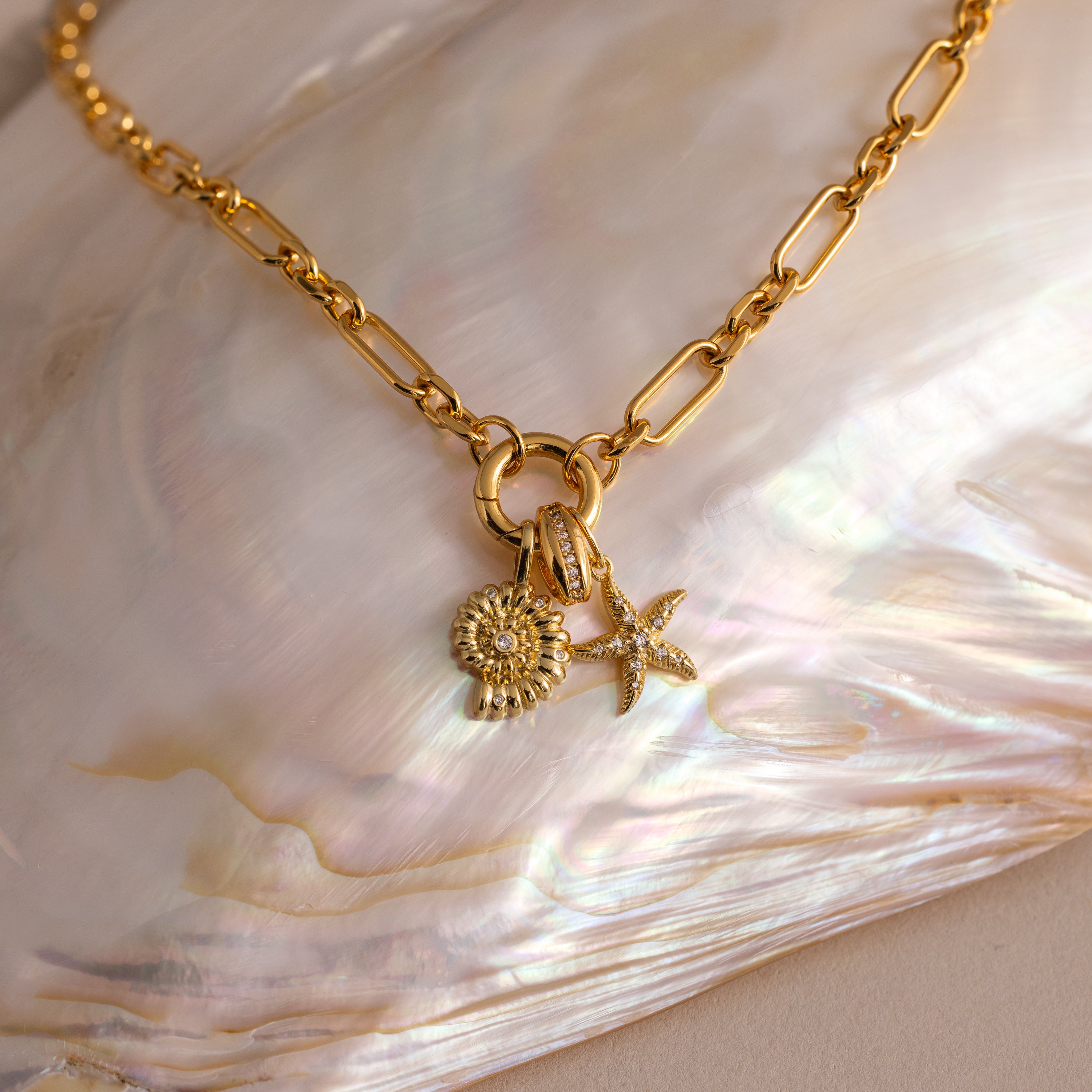 The Rolo Circle Figaro Necklace in 18K Gold features daisy and starfish charms, a statement front clasp, and is displayed on an iridescent shell for a modern update to the classic link style.