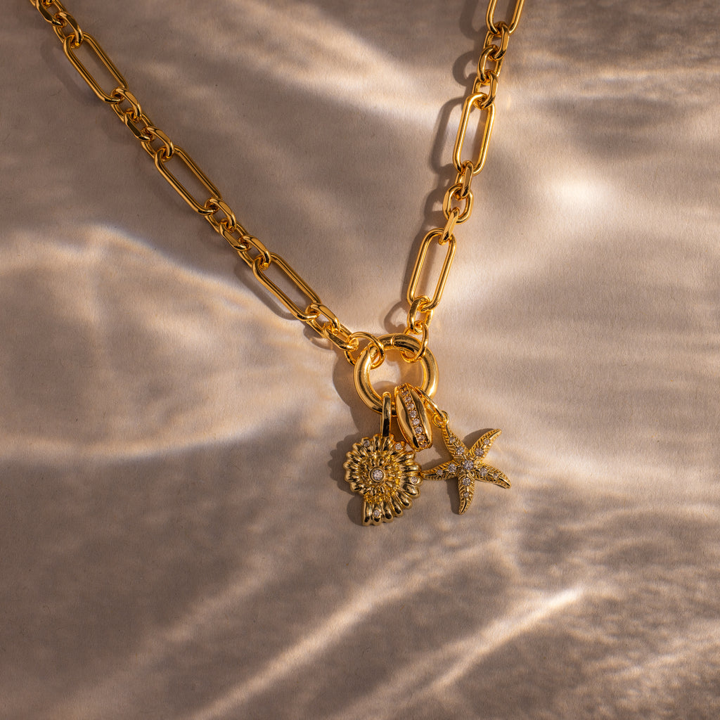 The Pave Dome Charm in 18K Gold adds subtle sparkle with its sun, shell, and starfish-inspired design, perfect for a gold chain necklace on a textured, sunlit surface.