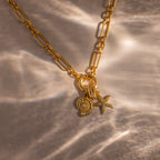 The Pave Dome Charm gold chain necklace features a stack of sun, shell, and starfish charms on a textured beige surface—an ideal accessory for fans of delicate jewelry.