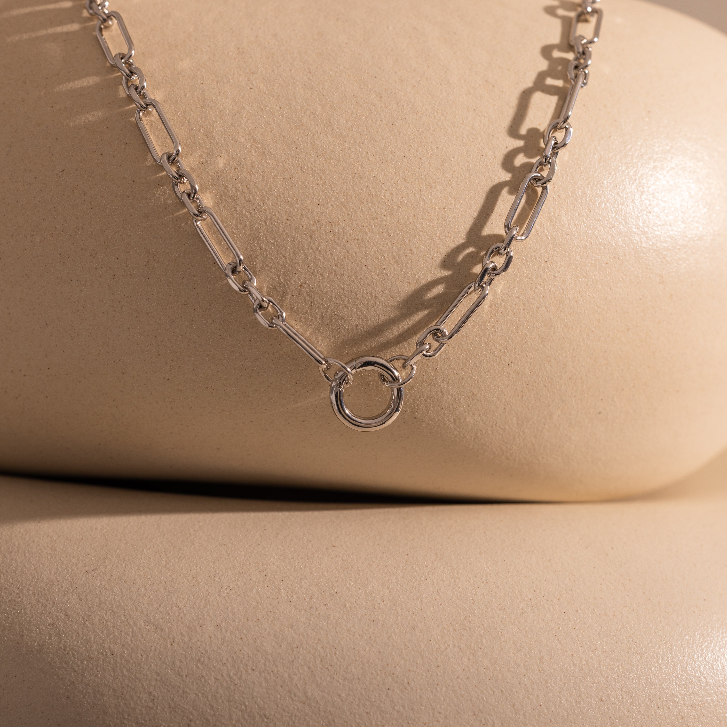 The Rolo Circle Figaro Necklace in Silver, featuring an open circle pendant and shown on smooth beige surfaces, creates a striking statement piece.