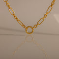 The Rolo Circle Figaro Necklace, a gold chain with a circular pendant, is reflected on a smooth beige surface for a look of effortless elegance.