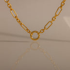 The Rolo Circle Figaro Necklace, a gold chain with a circular pendant, is reflected on a smooth beige surface for a look of effortless elegance.