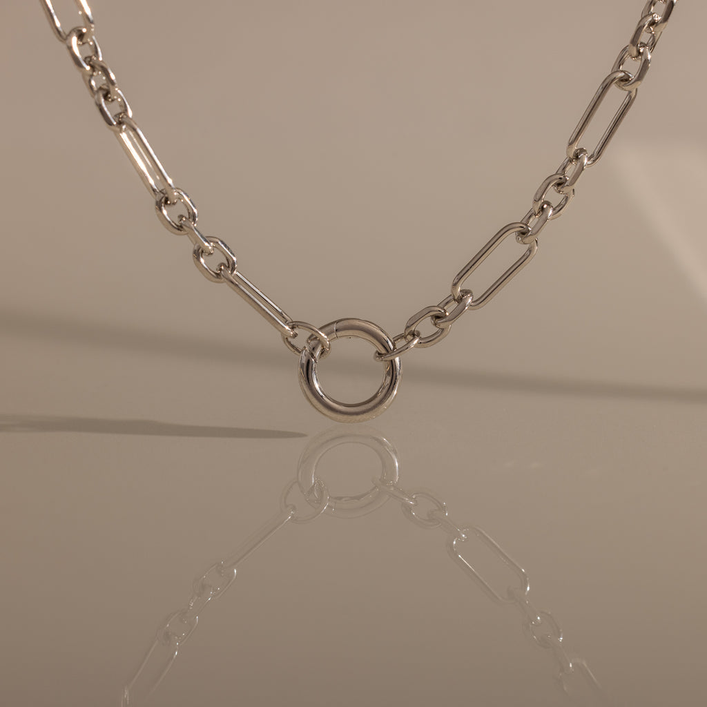 The Rolo Circle Figaro Necklace in Silver features bold links and a circular pendant, beautifully reflected on a smooth surface—a true statement piece for any jewelry collection.