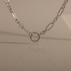 The Rolo Circle Figaro Necklace in Silver features bold links and a circular pendant, beautifully reflected on a smooth surface—a true statement piece for any jewelry collection.