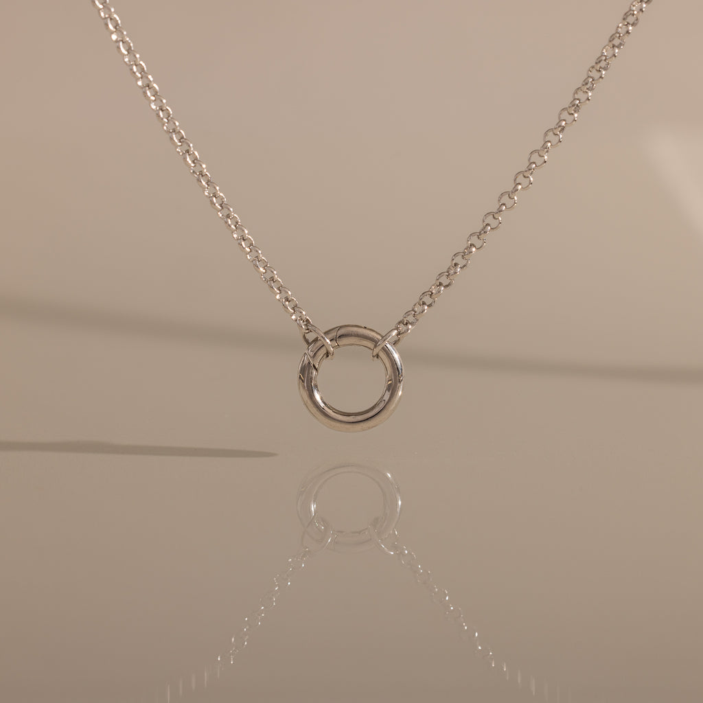 The Cable Circle Charm Necklace in Silver features a simple silver chain with a small circular pendant, elegantly reflected on a smooth surface.